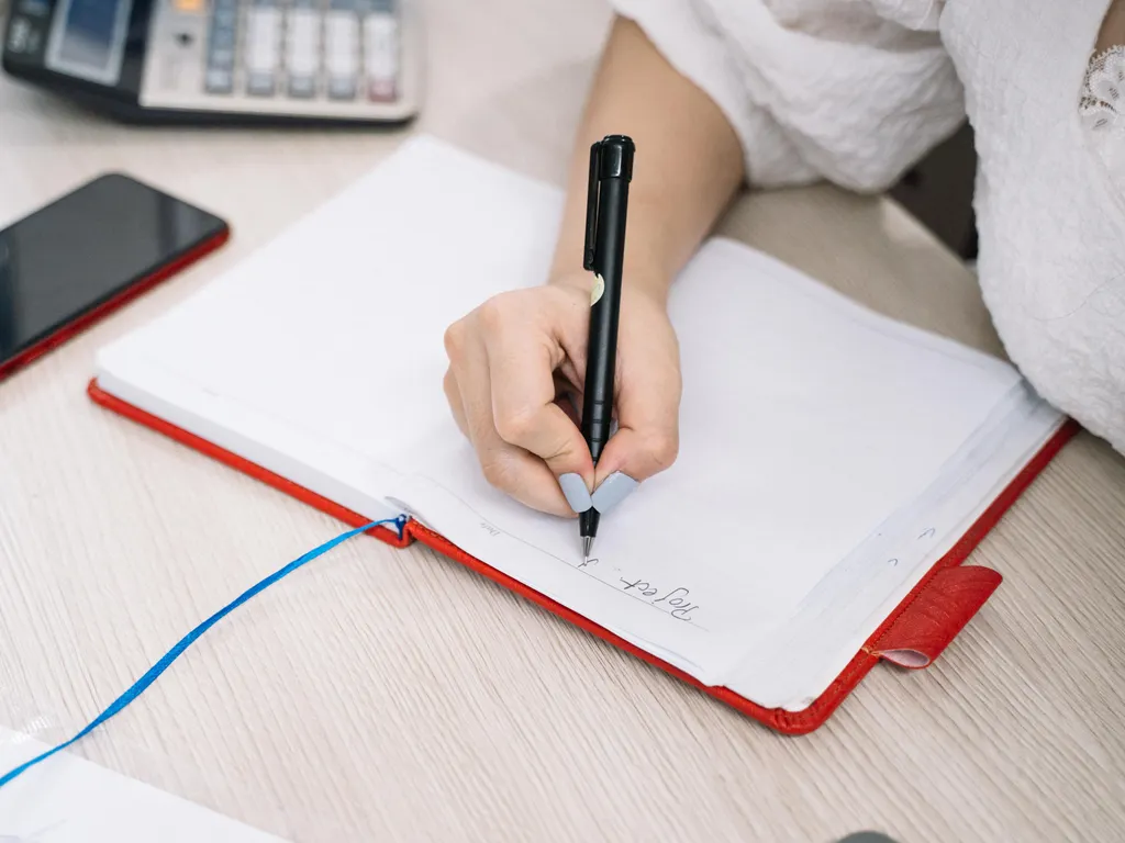Masada açık defter, telefon ve hesap makinesiyle aylık gider notu alan el